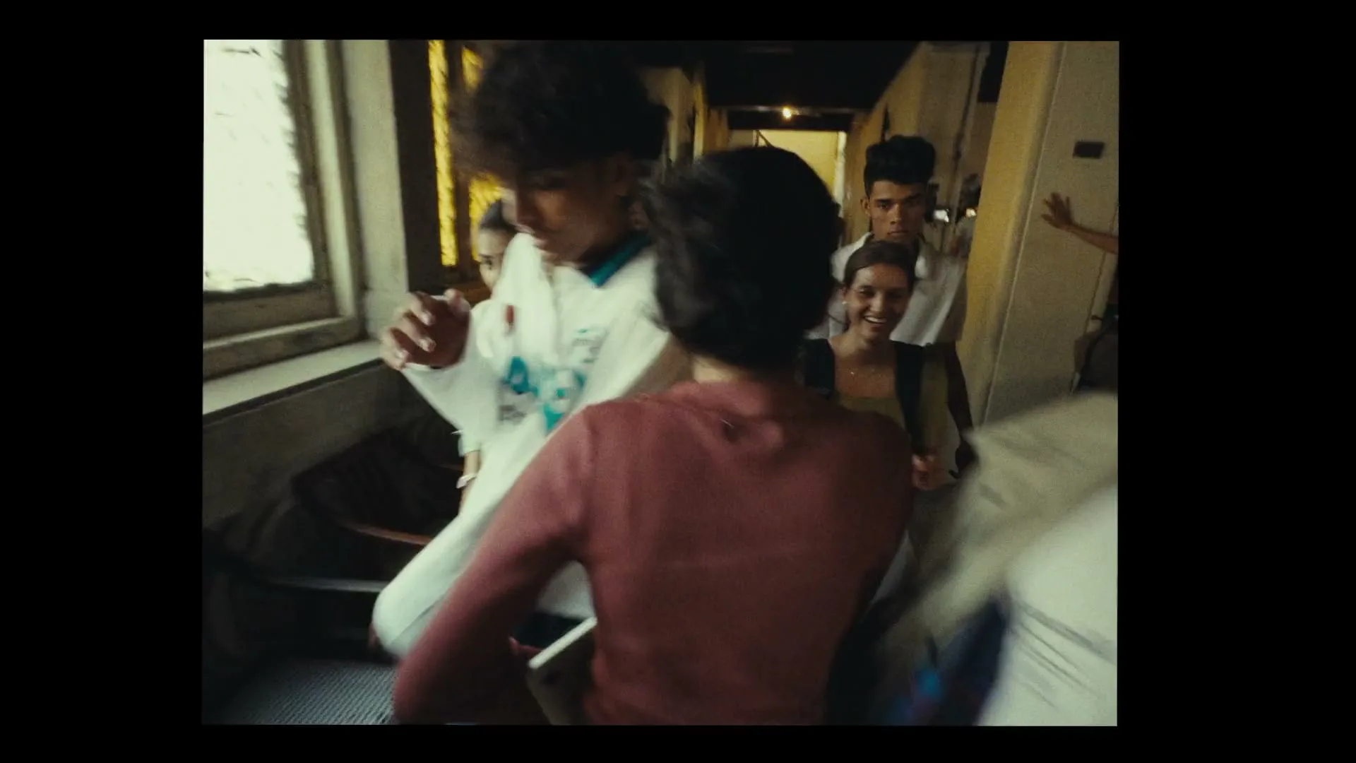 A diverse group of young people walk through a narrow, sunlit hallway, some smiling, in a dynamic, handheld shot.