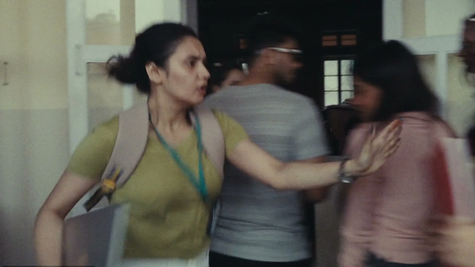 A young woman with a backpack and laptop pushes through a crowded hallway, looking stressed and trying to make her way through the people.