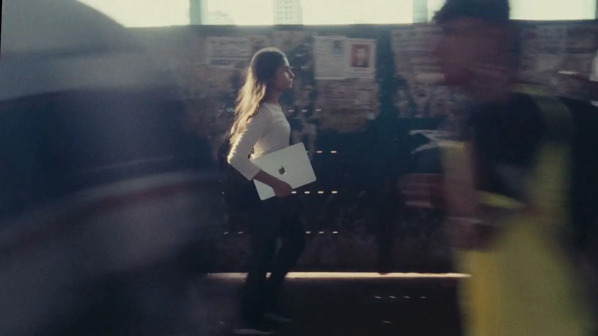 A young woman in a white top and dark pants walks through a busy, sunlit interior space, carrying a silver laptop.