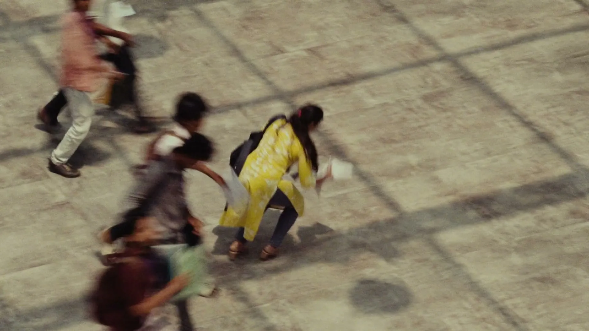 A group of students, including a woman in a yellow dress, scramble to pick up scattered papers on a tiled floor in an exterior setting.