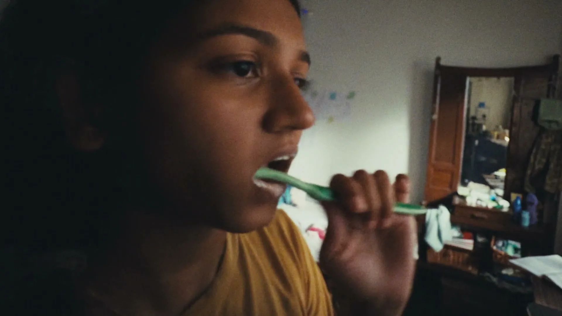 A young woman with dark hair and a yellow shirt brushes her teeth in a dimly lit room, with a mirror and dresser in the background.