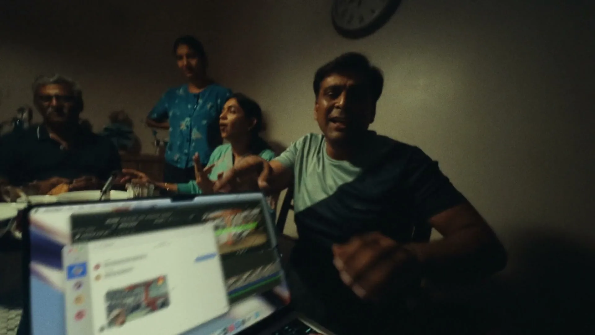 A man in a t-shirt sits at a table with family, gesturing while looking at a laptop screen, in a dimly lit interior.