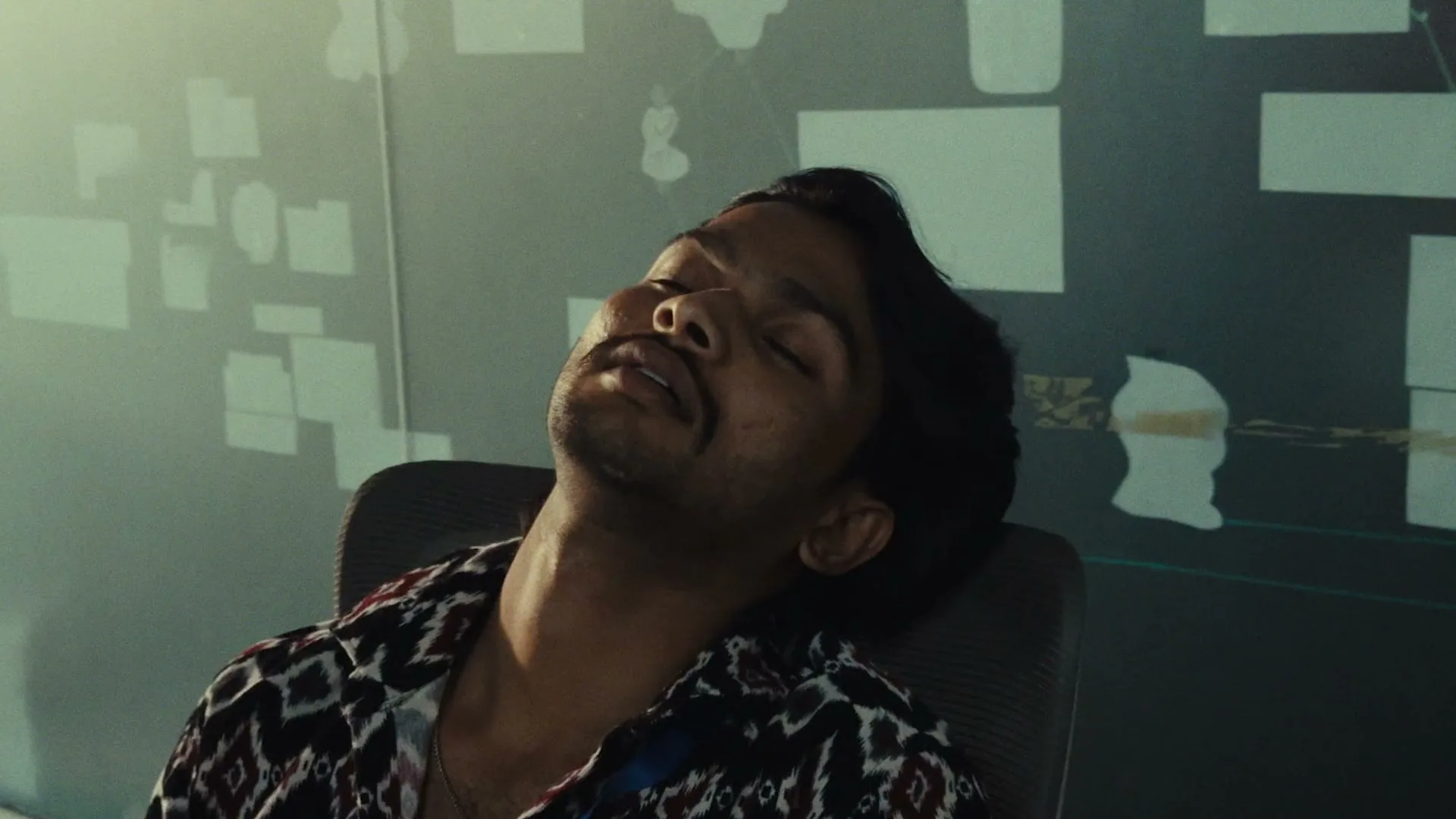 A man with a mustache and dark hair, wearing a patterned shirt, is sleeping in an office chair, then is startled awake by someone off-screen.