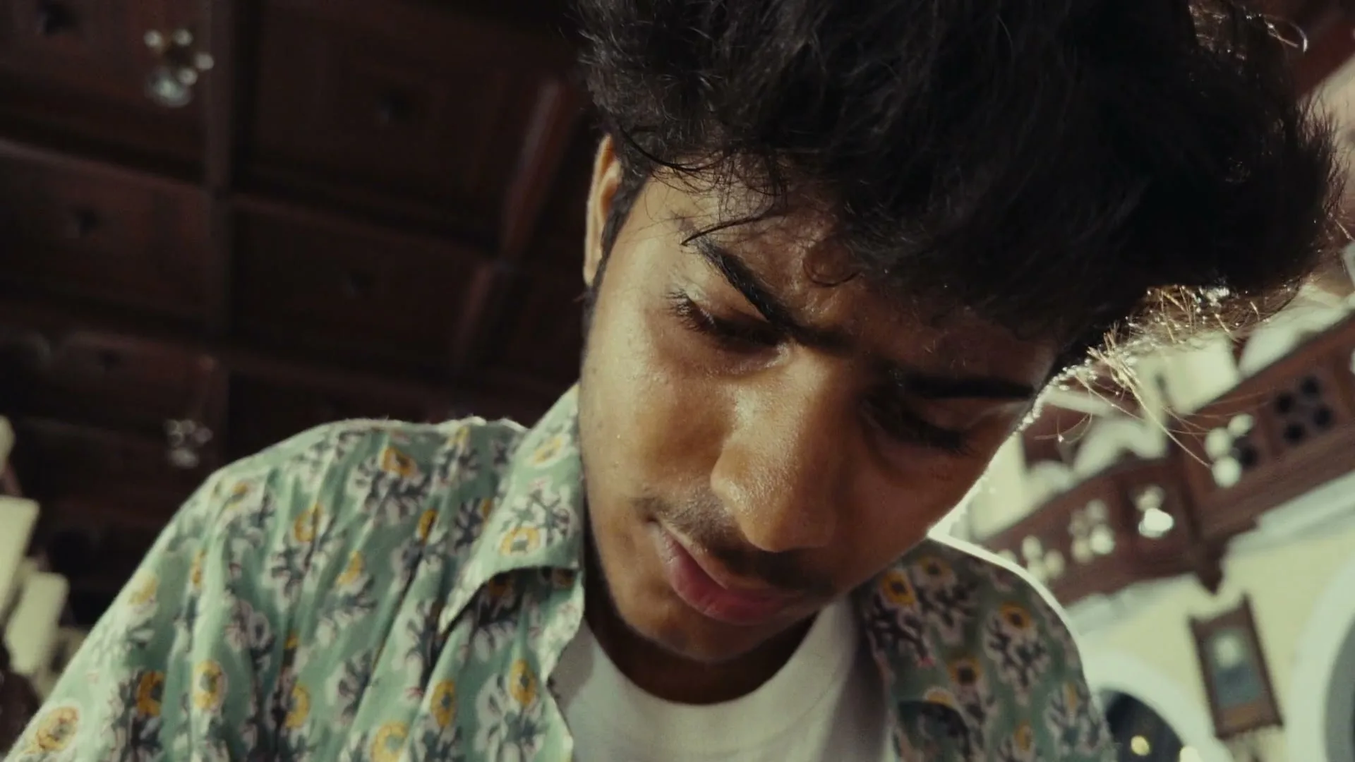 A young man with dark, curly hair and a patterned shirt looks down intently, focused on something below the frame, in an ornate interior setting.