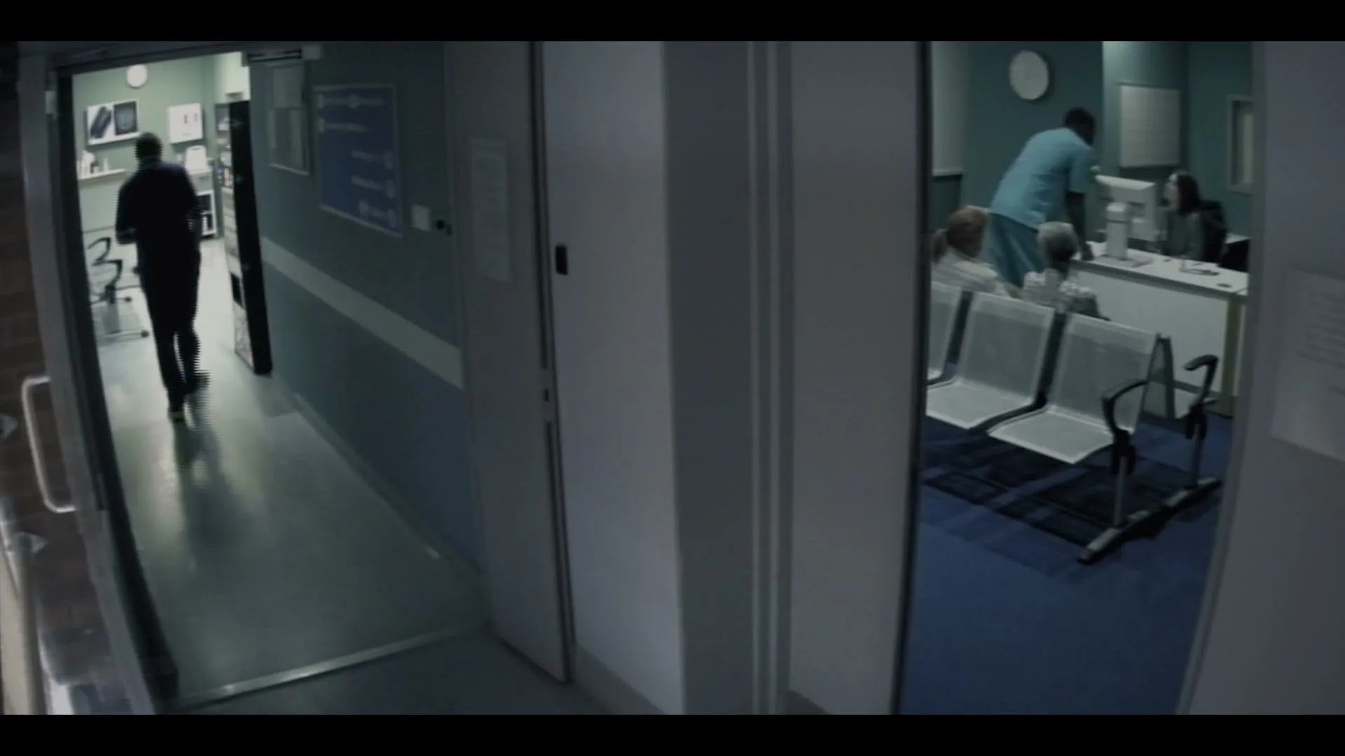A male doctor in scrubs walks down a hospital corridor while patients wait in a waiting room.