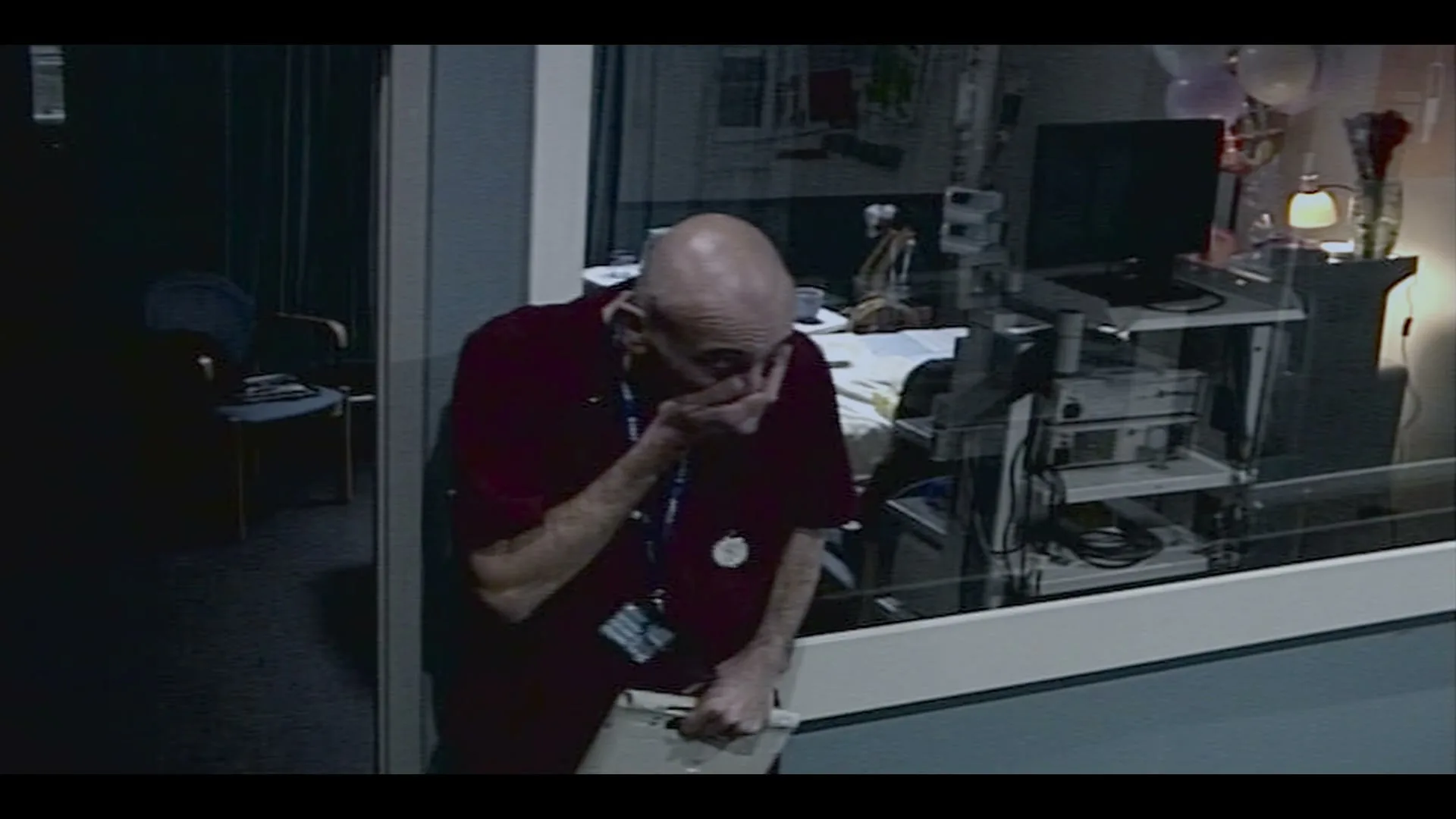 A bald male doctor in red scrubs cries, covering his mouth, while holding a clipboard in a hospital hallway next to a patient's room.