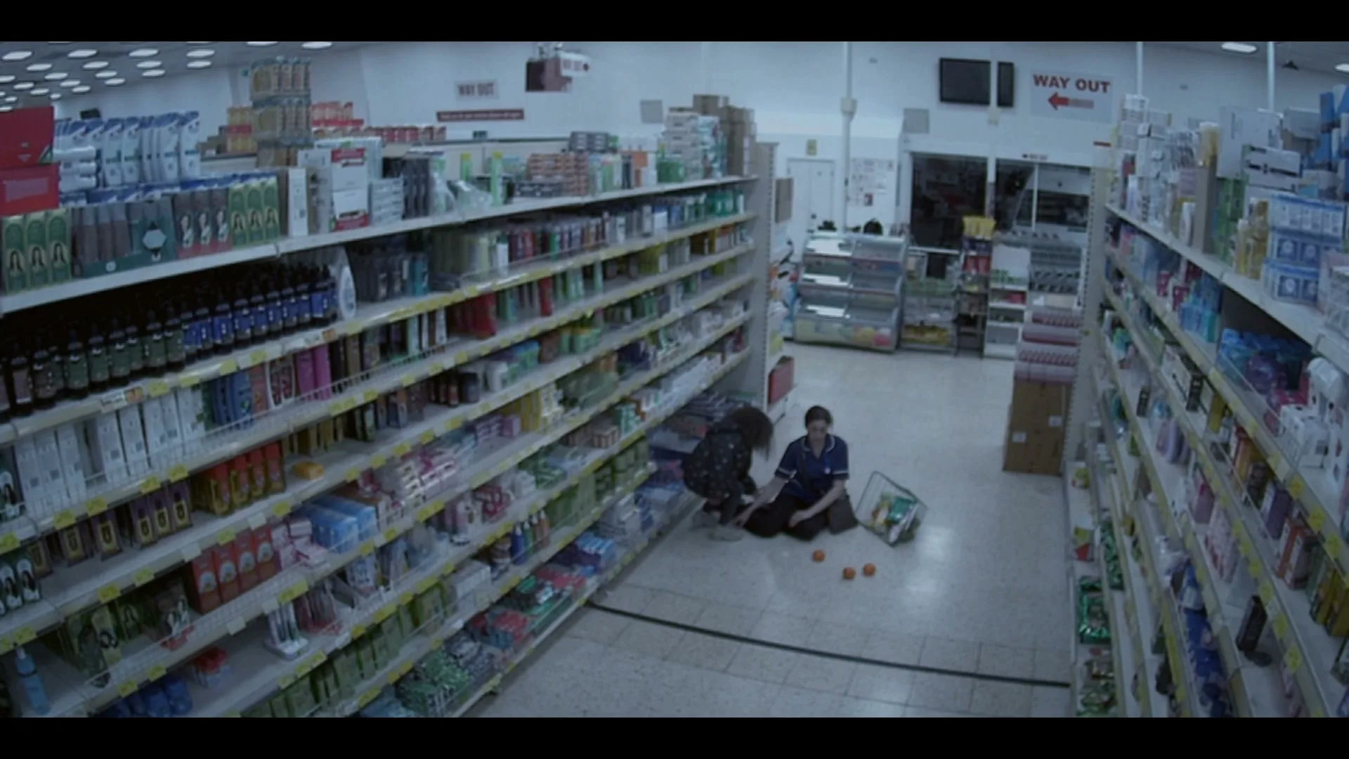 A young woman in a dark jacket helps a male nurse in scrubs pick up oranges that have fallen from his shopping basket in a brightly lit supermarket aisle.