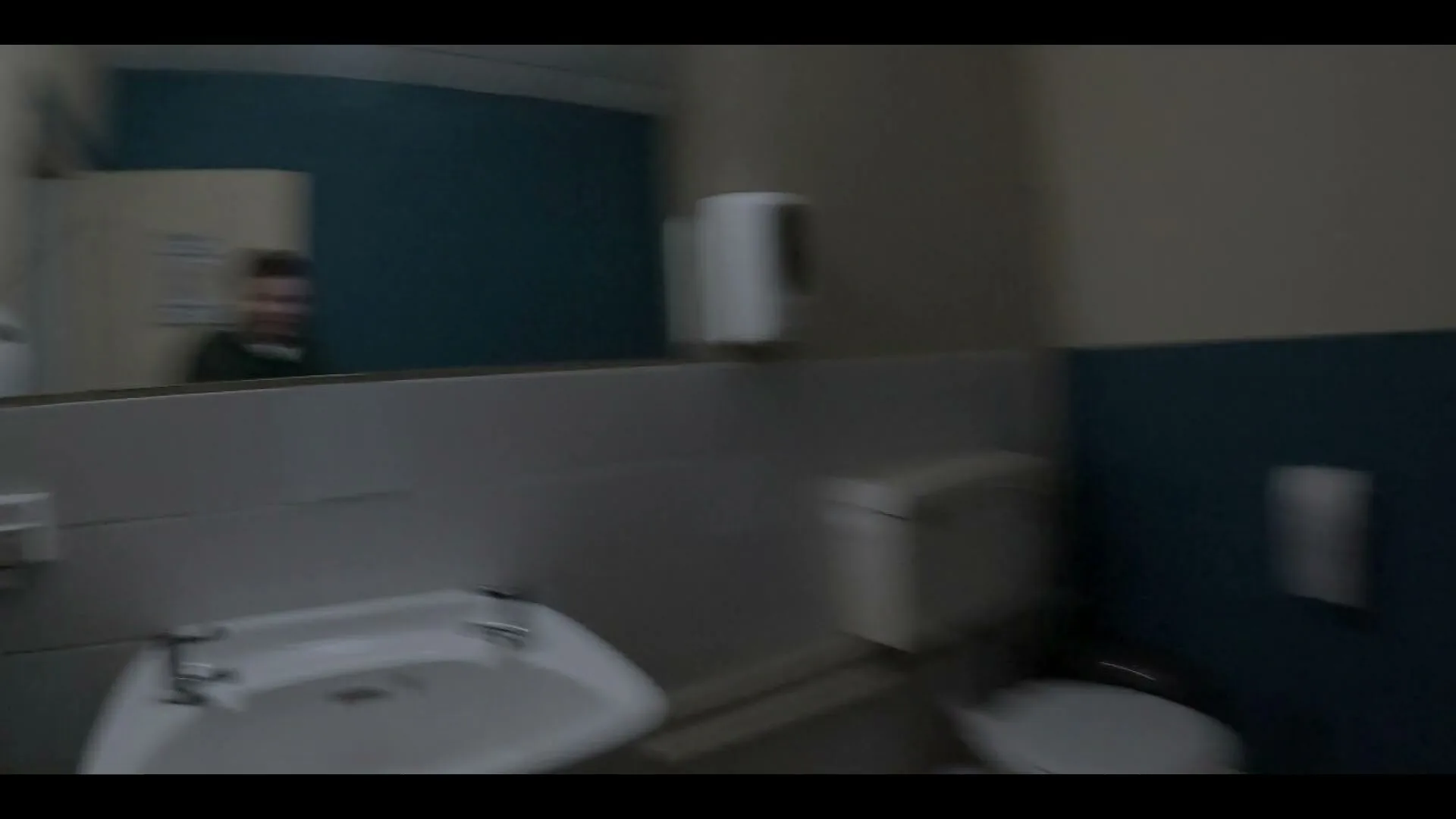 A person in blue gloves, covered in blood, looks at their hands in a hospital bathroom mirror, while another person watches in the reflection.