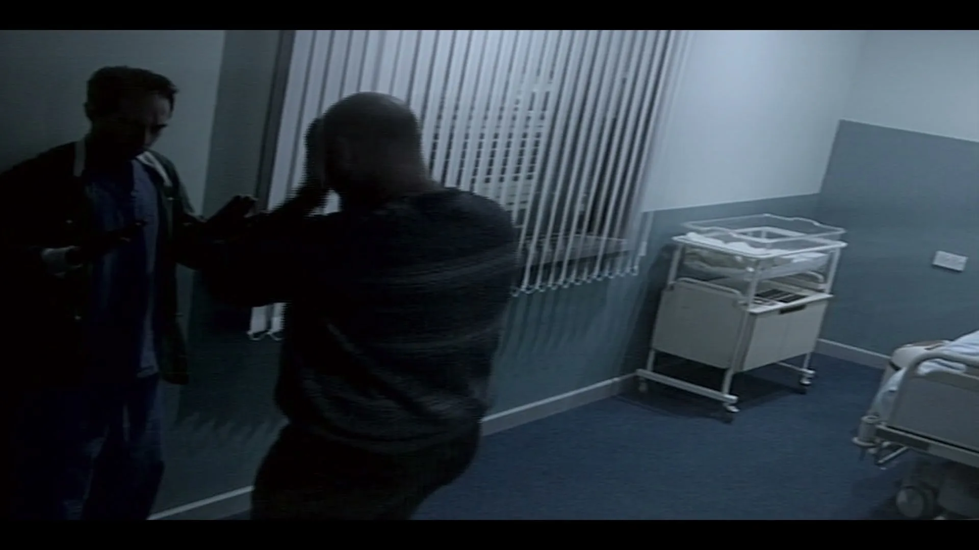 Two men, one in scrubs and one in a striped shirt, stand in a dimly lit hospital room with a baby bassinet and bed.