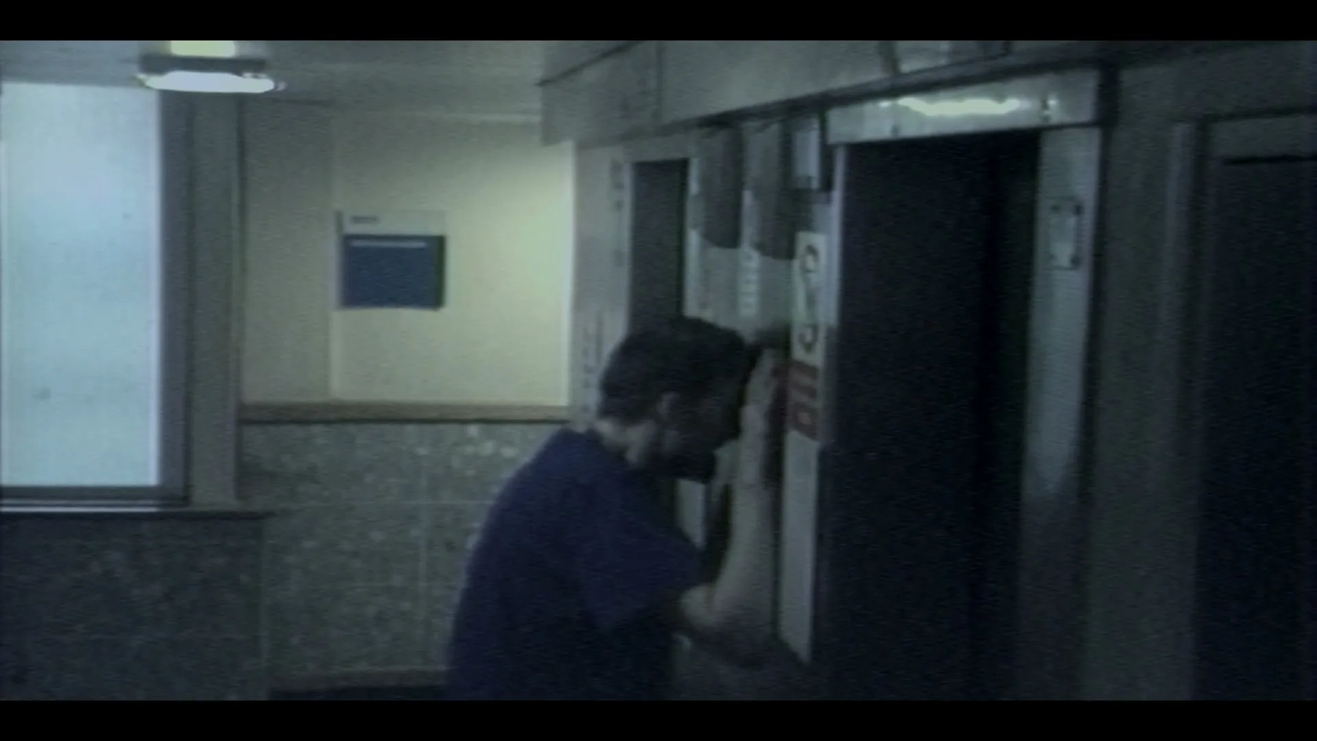A male healthcare worker in blue scrubs leans his head against a hospital elevator door, looking exhausted and overwhelmed in a dimly lit hallway.