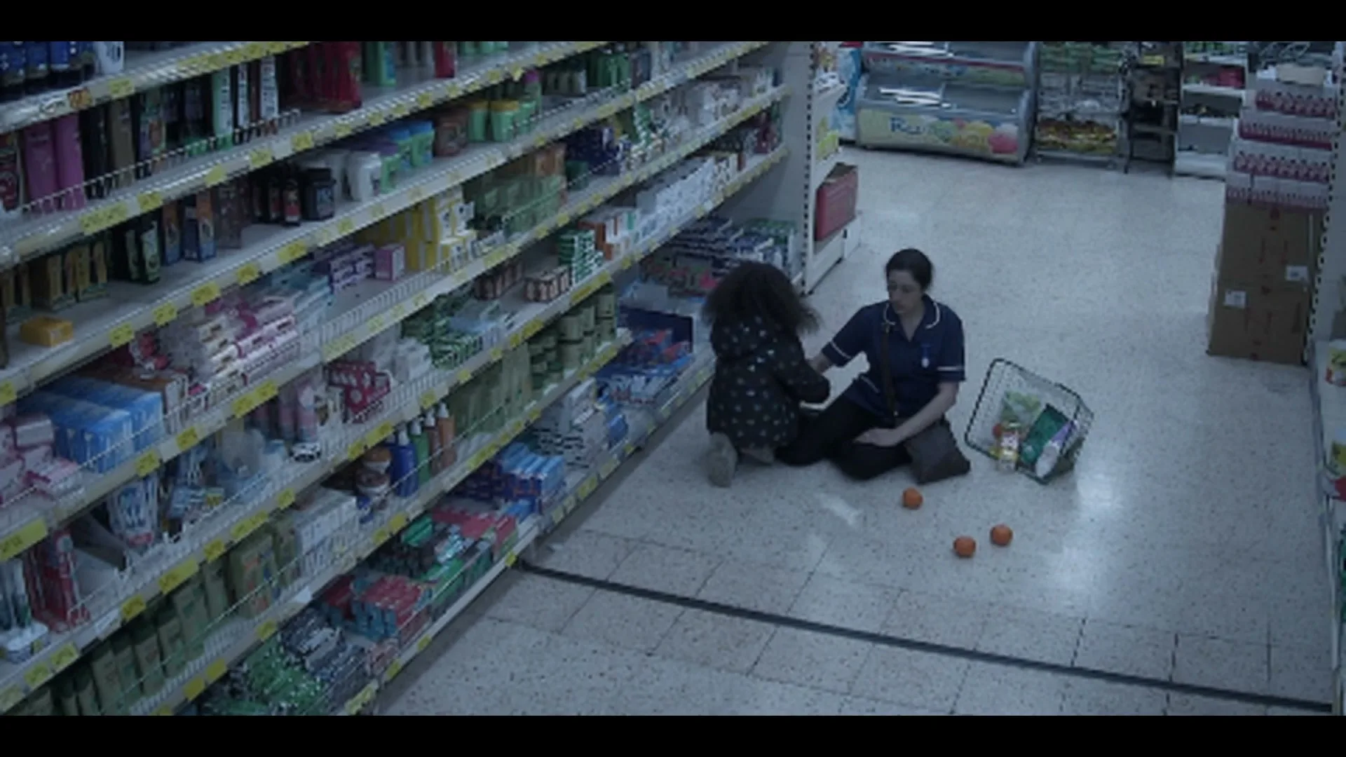 A female nurse in blue scrubs kneels on the floor of a grocery store aisle, comforting a distressed woman with curly hair.