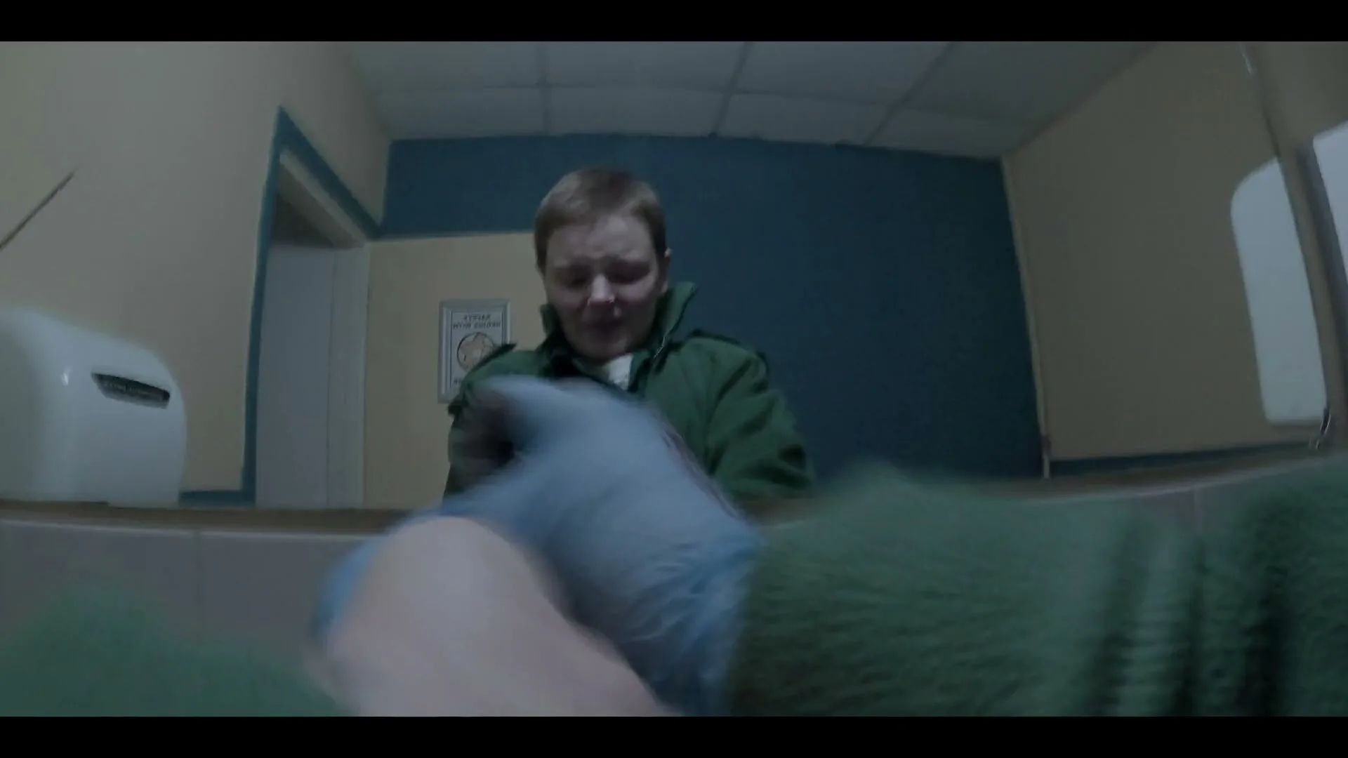 A person in a green jacket and blue gloves is seen from a low angle, looking down at something out of frame in a hospital bathroom.