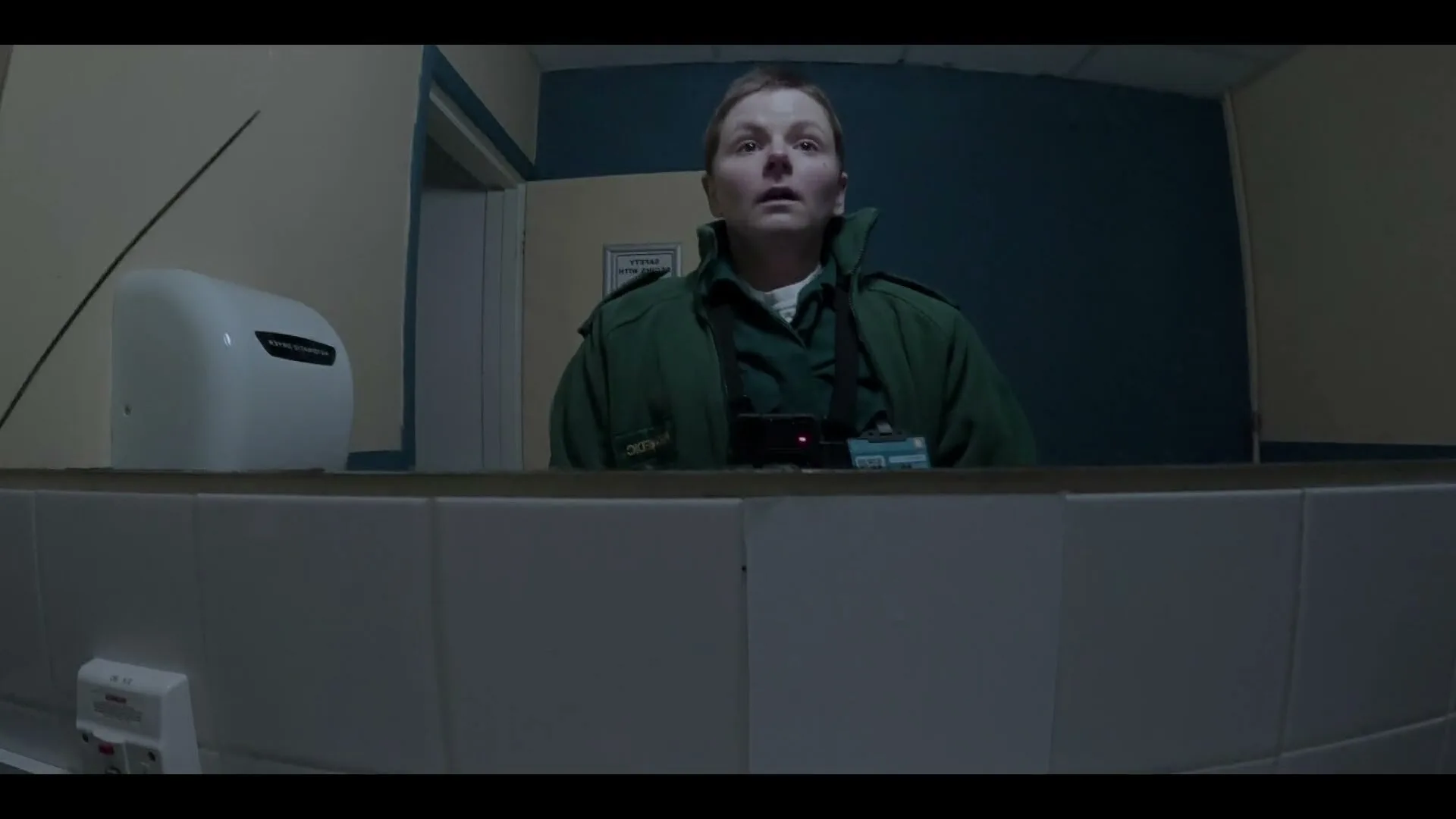 A female paramedic in a green uniform looks at her reflection in a mirror in a dimly lit public restroom, then wipes the mirror with her hand.