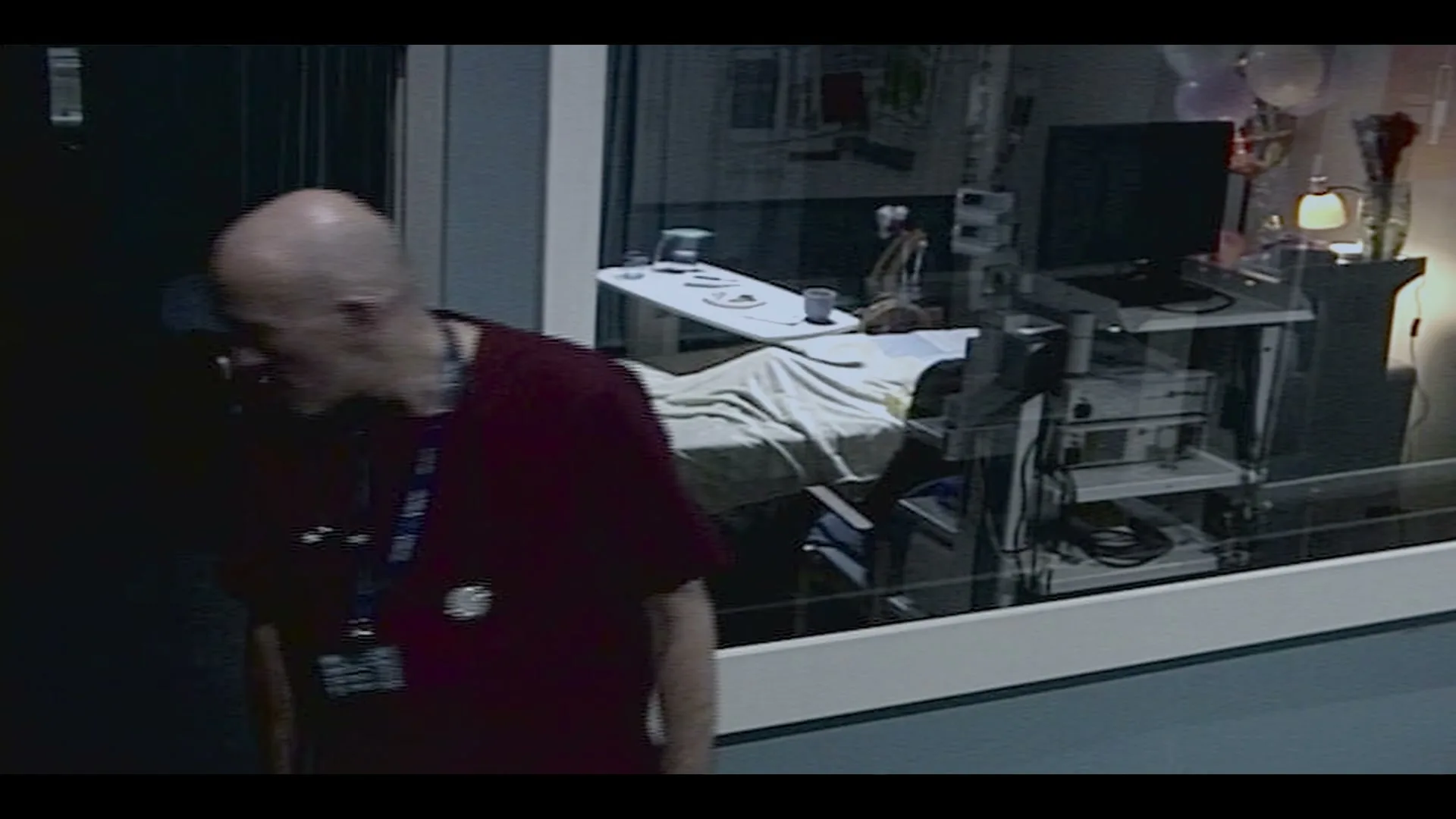 A bald male doctor in maroon scrubs and a stethoscope walks past a hospital room with a bed and medical equipment, looking tired.