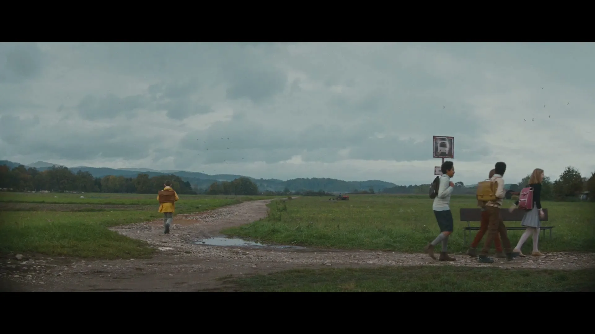 A group of children walk along a dirt path next to a field, as a bus drives past them on a cloudy day.