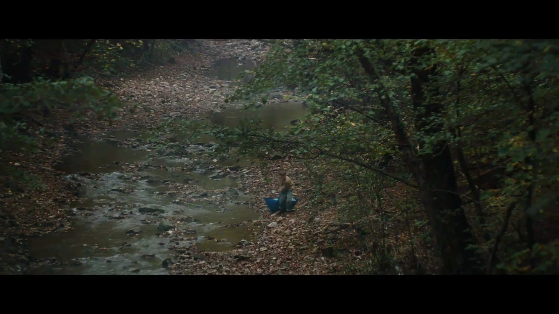 A young man in a beige shirt and blue pants is seen from above, standing next to a blue bucket in a rocky stream bed surrounded by trees.