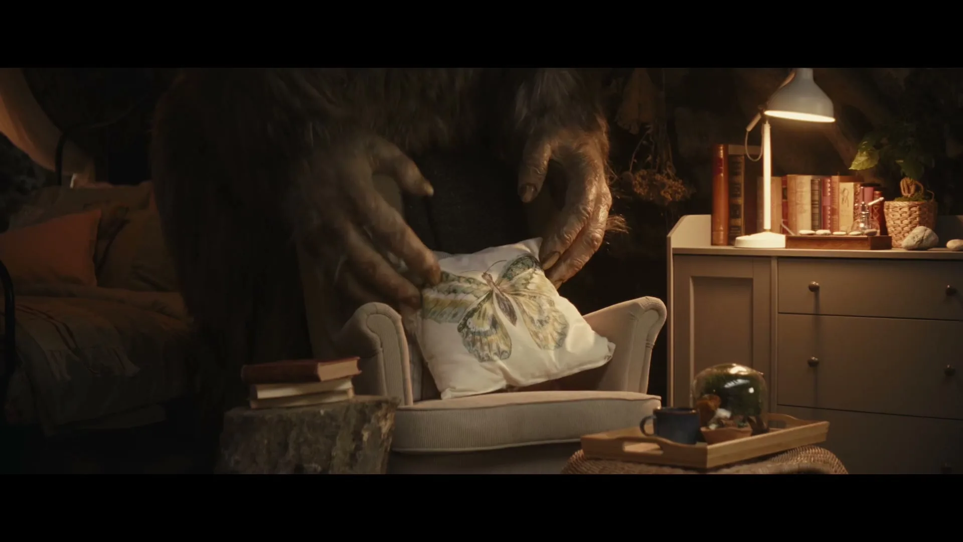 A large, furry troll's hands gently place a decorative pillow with a butterfly design onto a small armchair in a cozy, dimly lit room.