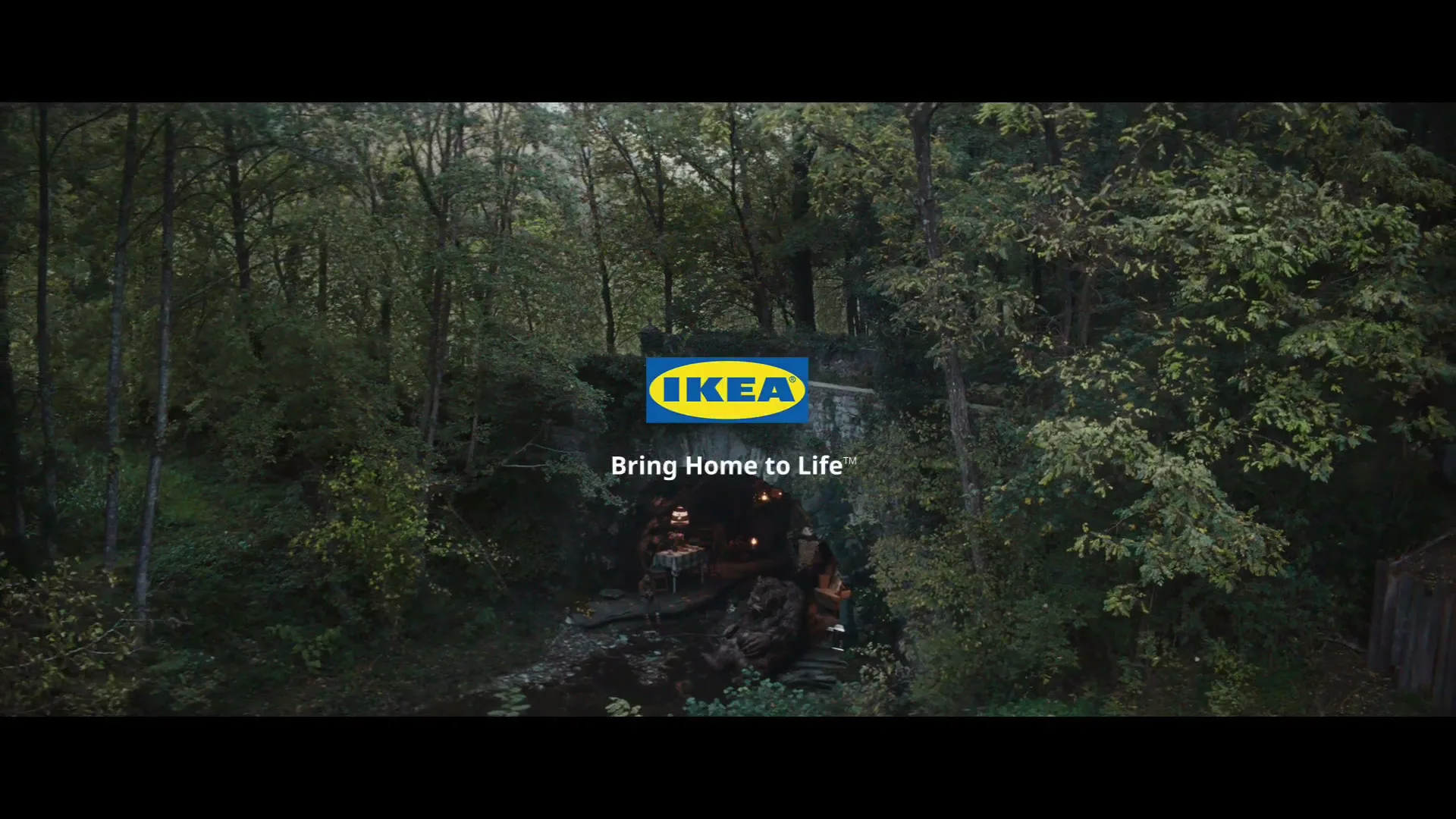 A wide shot of a lush green forest with a stone bridge over a stream, revealing a cozy, candlelit interior beneath the archway, with the IKEA logo and tagline appearing over the scene.