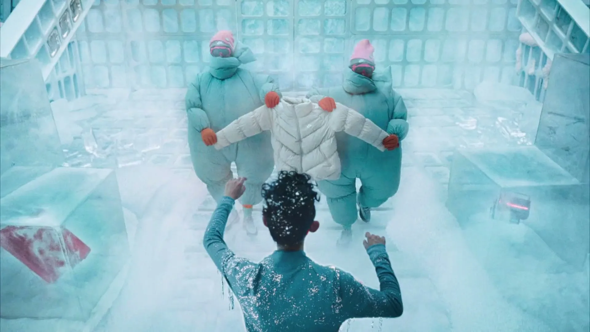 Two people in puffy suits present a white puffer jacket to a person covered in ice in a futuristic ice room.