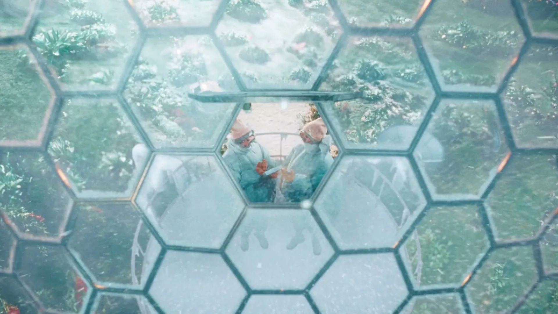 Two people in winter gear are seen through a hexagonal glass dome, looking out at a snowy landscape with green plants.