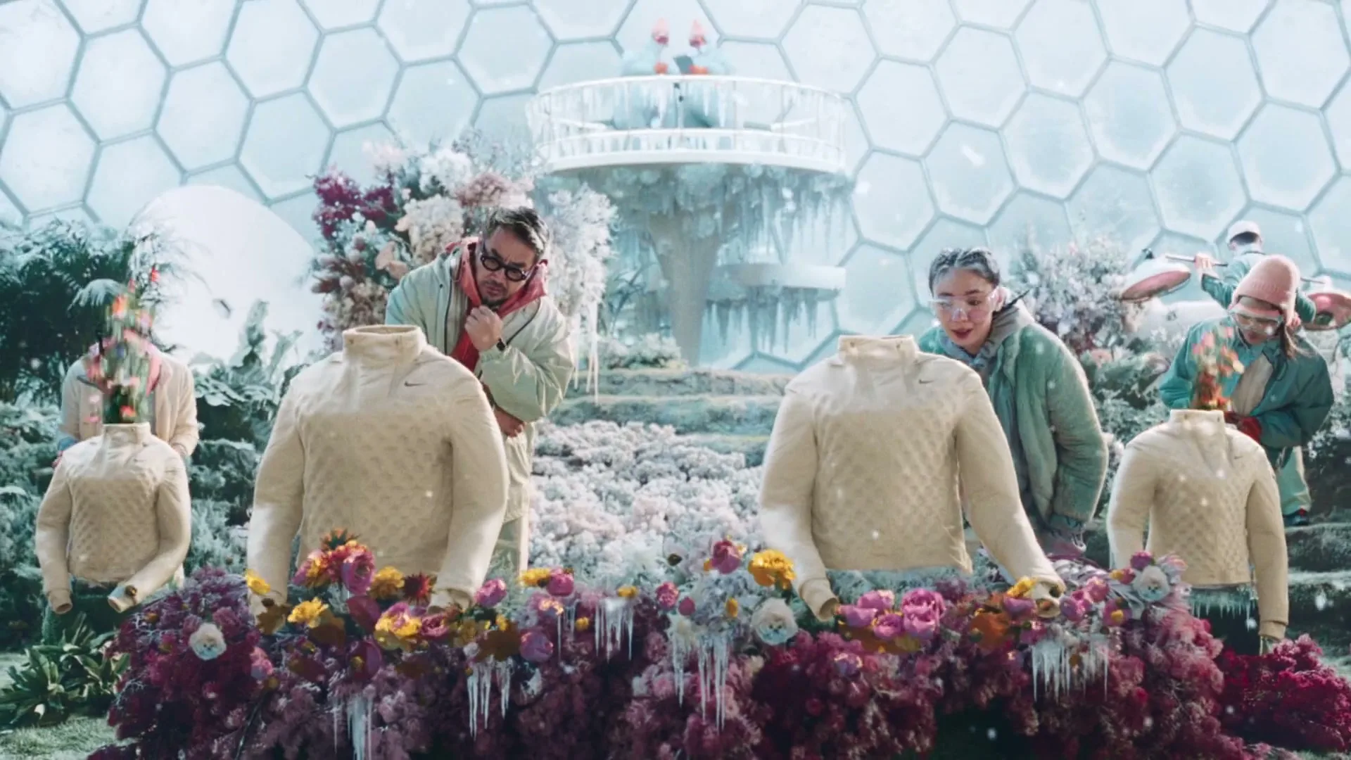 People in winter clothes watch as flowers sprout from headless mannequins wearing cream-colored sweaters in a futuristic, hexagonal-paneled greenhouse.