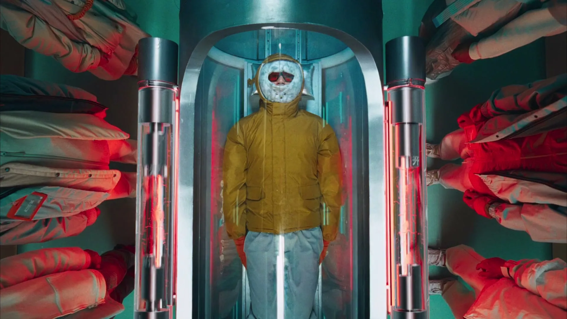 A person in a yellow jacket, white pants, and a hockey mask stands in a futuristic chamber, surrounded by hanging clothes.