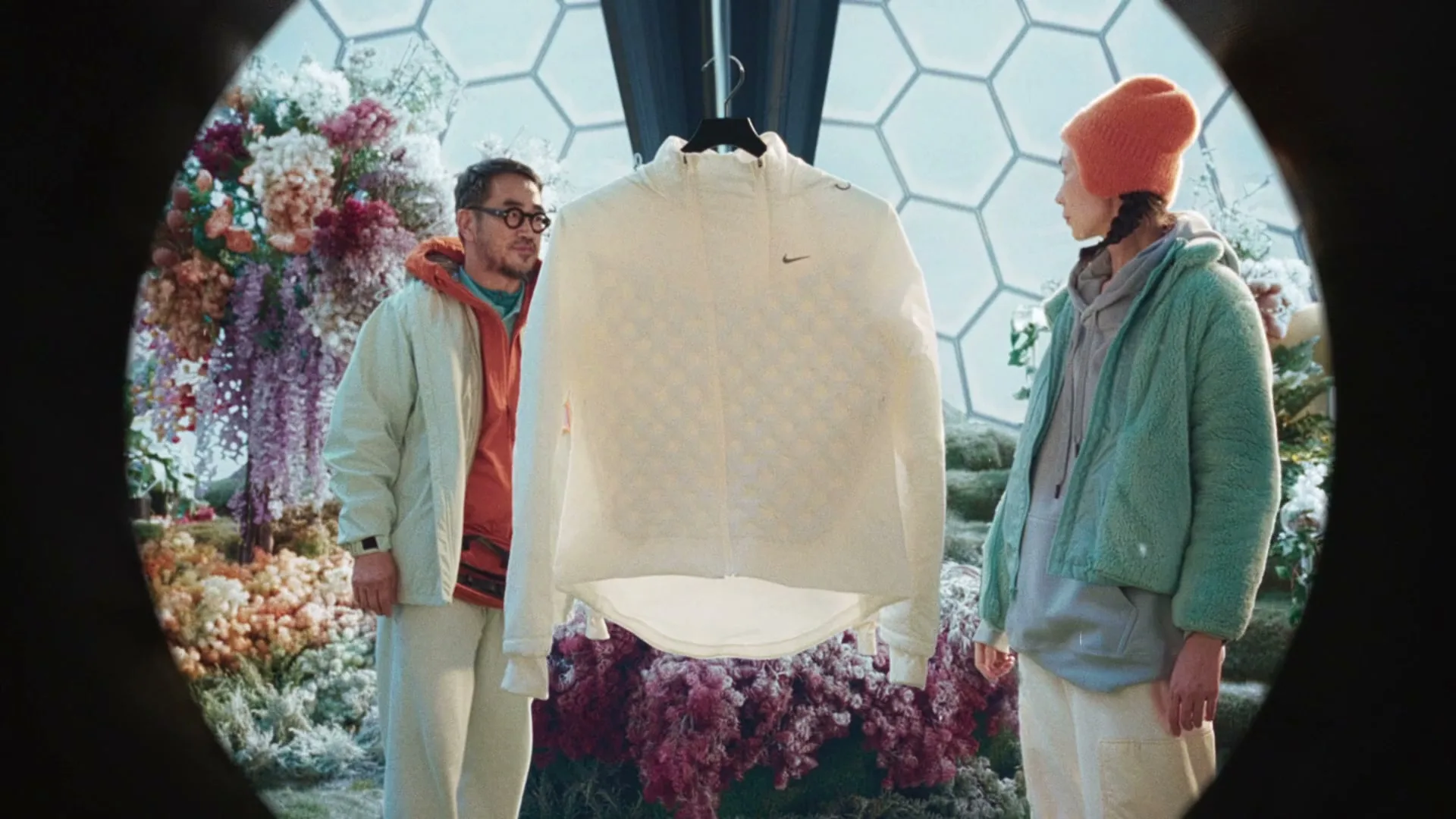 A man and a woman stand on either side of a white Nike jacket, which hangs suspended in a lush, floral, futuristic greenhouse.