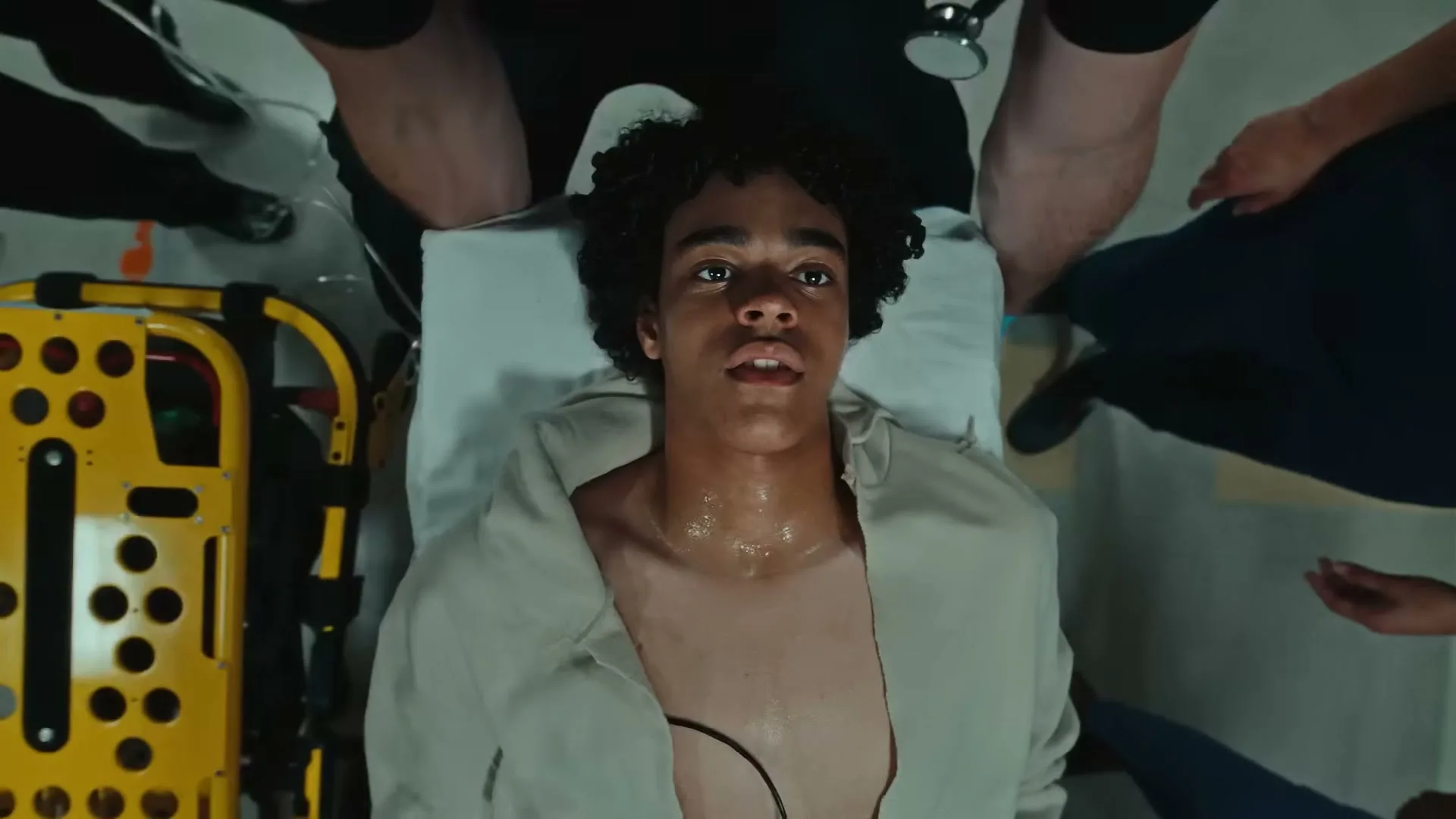 A young male patient with curly hair lies on a stretcher, looking up, surrounded by medical personnel in a hospital setting.