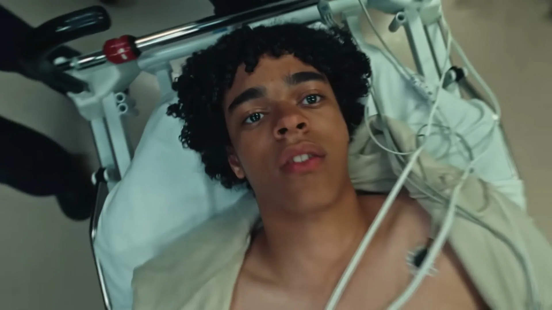 A young male patient with curly hair lies on a hospital gurney, connected to medical equipment, looking up as he is wheeled through a hospital corridor.