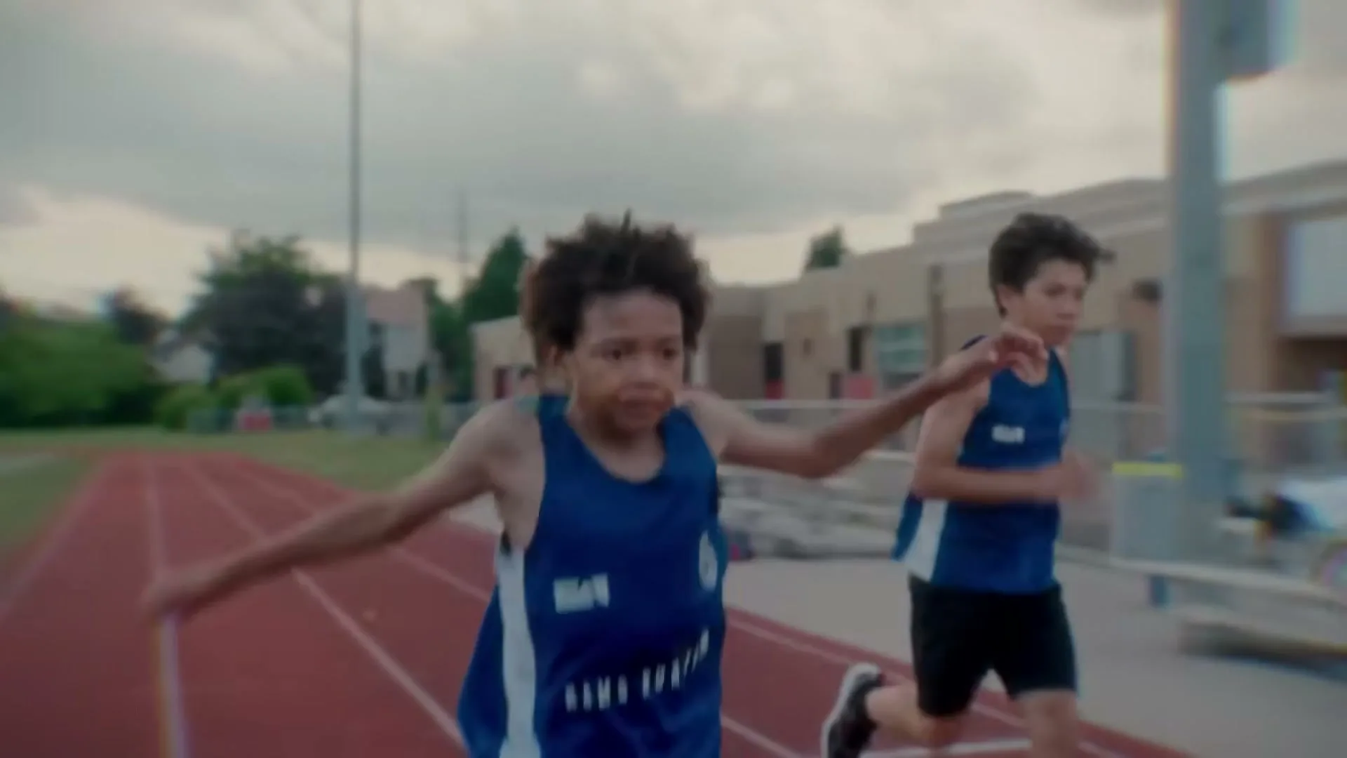Three young boys in blue track uniforms run on a red track at an outdoor school track, with a building in the background.