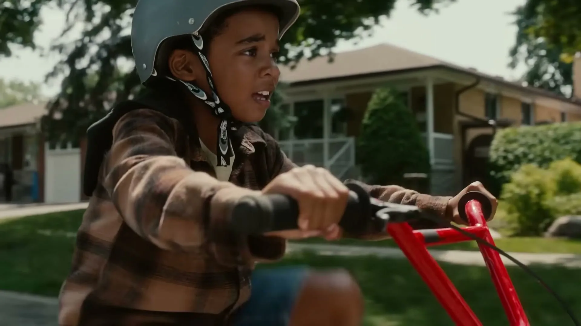 A young boy wearing a helmet and plaid shirt rides a red tricycle down a suburban street during the day, looking to his right with an open mouth.