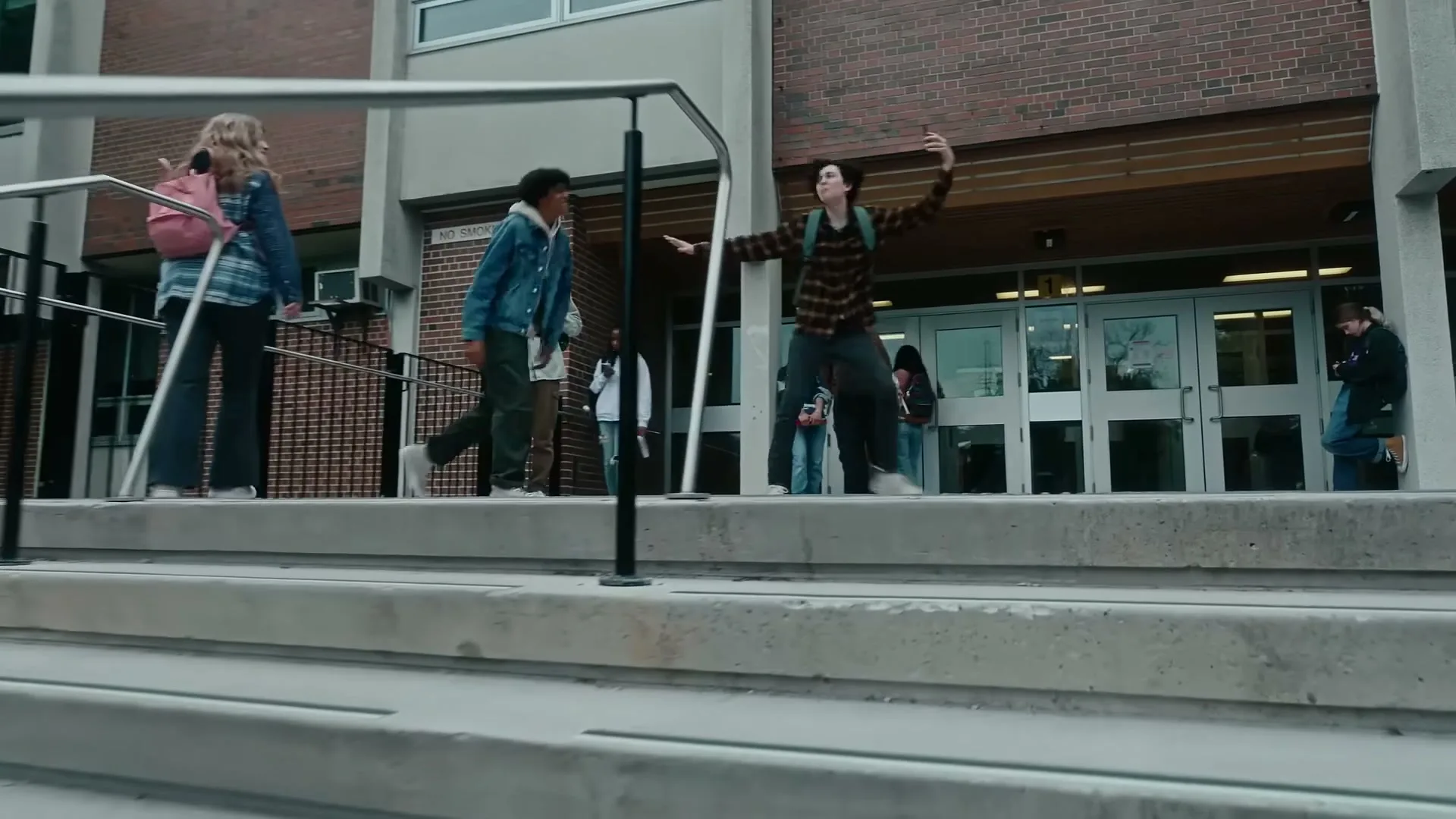 A diverse group of high school students walk out of a school building, with one male student in a plaid shirt dancing playfully on the steps.