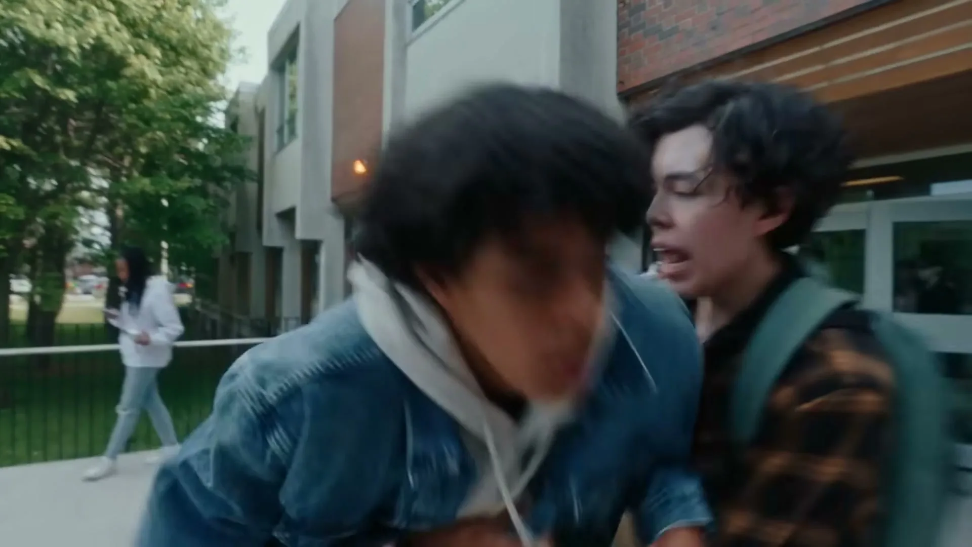 A male teen in a denim jacket clutches his chest and collapses to the ground outside a school building, as another male teen tries to help him.