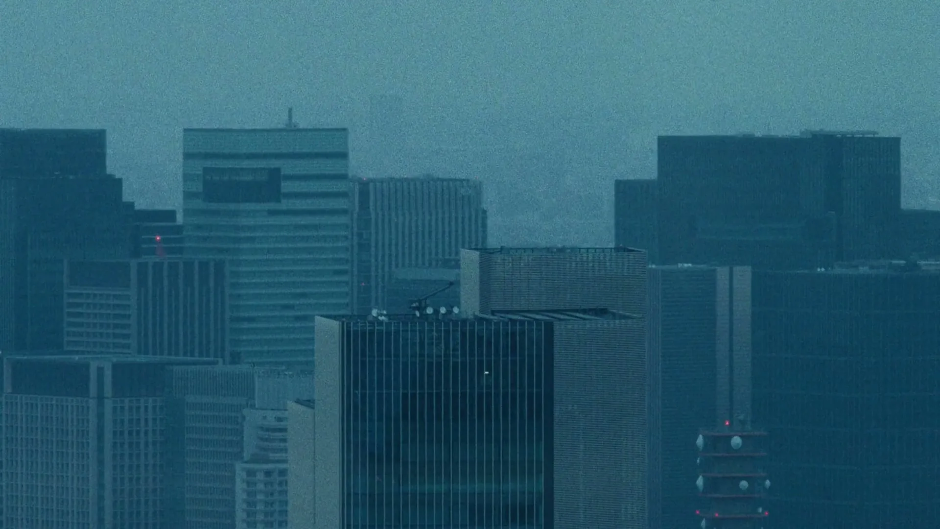 An extreme wide shot of a city skyline with tall buildings under a blue-grey sky, with Japanese and English text appearing on screen.