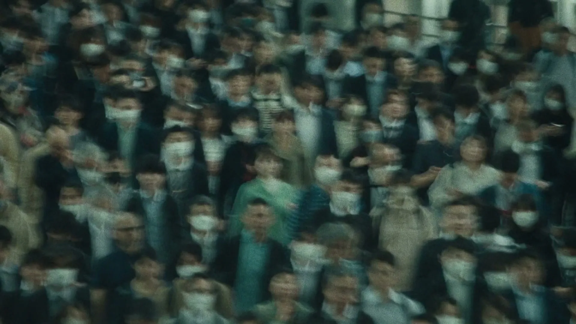 A blurred crowd of people, many wearing face masks, moves through a busy public space in an extreme wide shot.