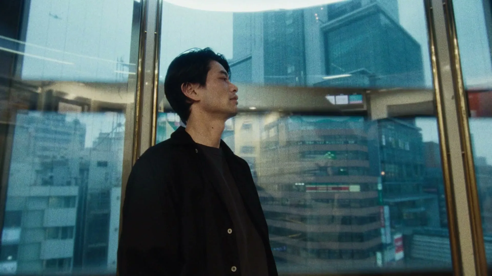 A man with dark hair and a black jacket looks out a window at a city skyline during the day.