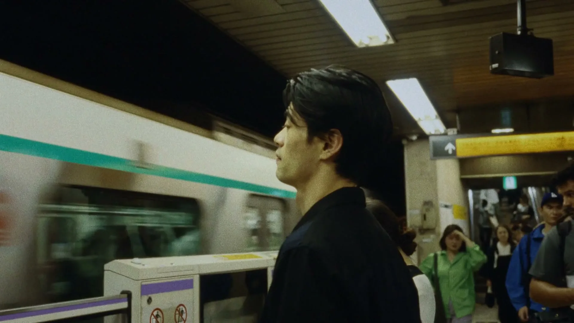 A young man with dark hair in a black shirt stands on a subway platform, looking forward as a train speeds past him.
