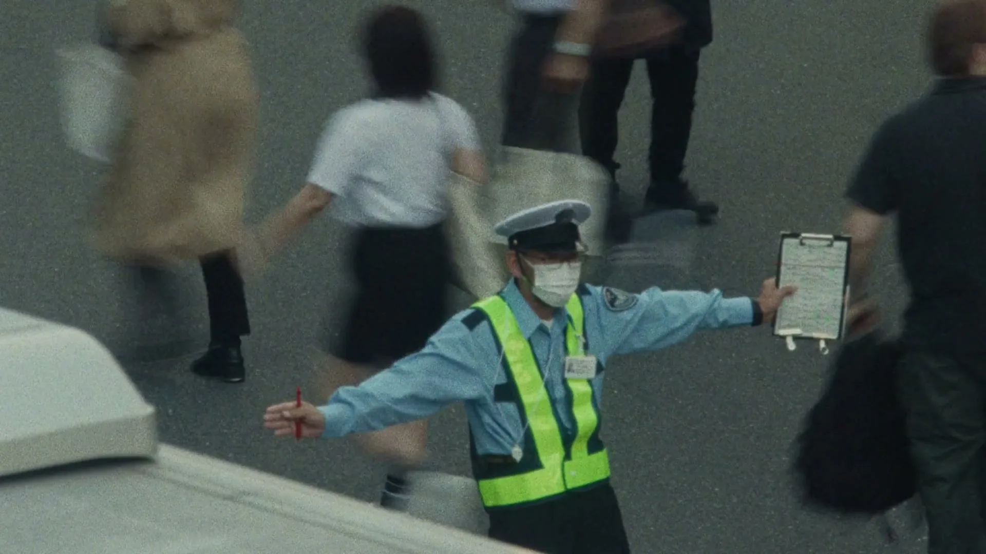 A traffic controller in a light blue uniform, white hat, and a neon yellow vest directs pedestrians in a busy city street during the day.