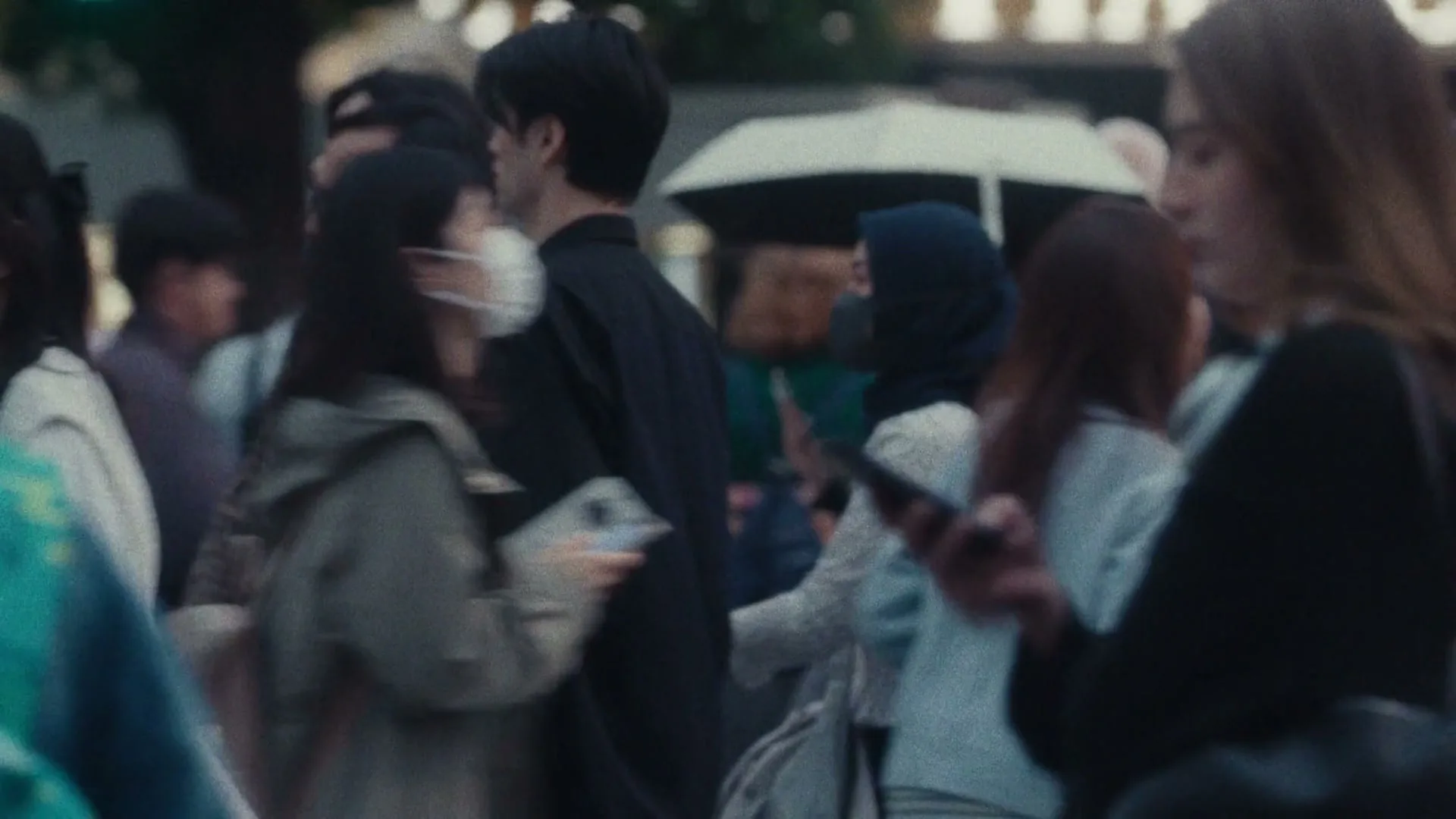 A crowd of diverse people walk on a city street, some looking at phones, with a blurred foreground element.