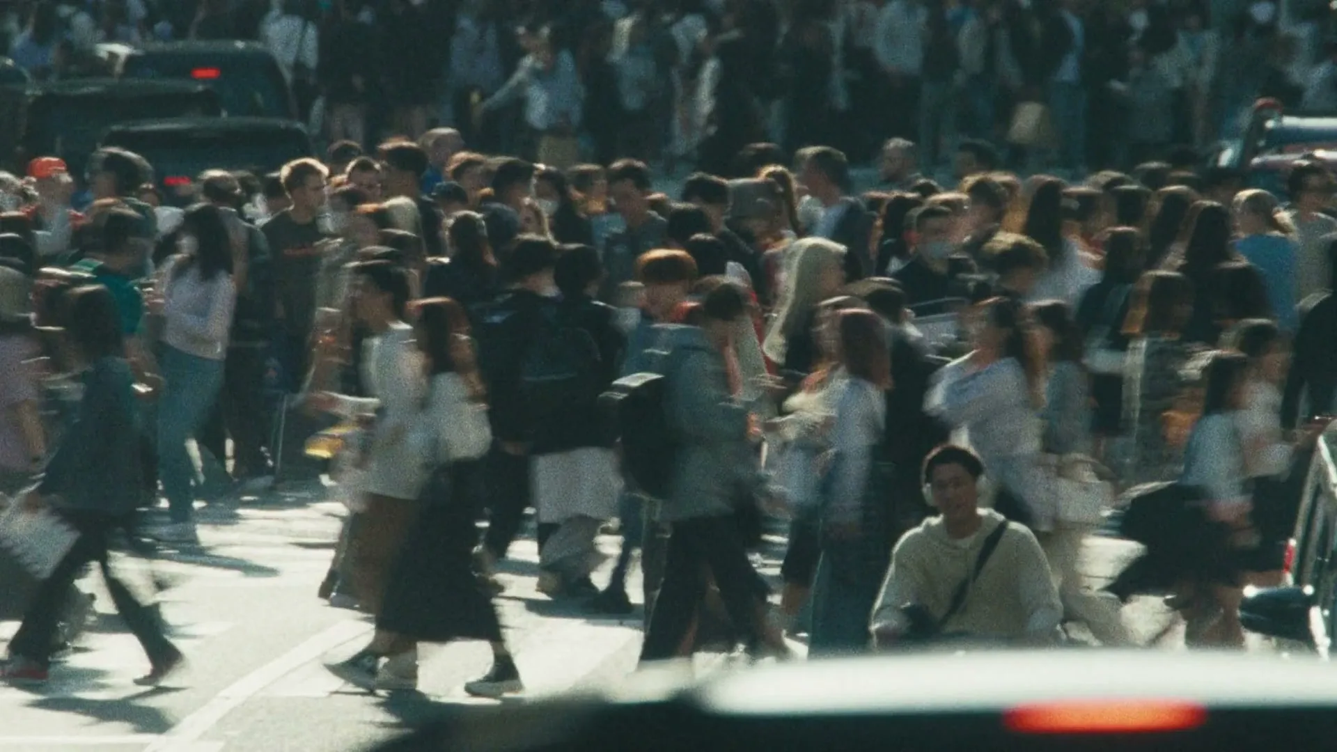 A crowd of people, mostly young adults, walk across a busy street in a city during the day.