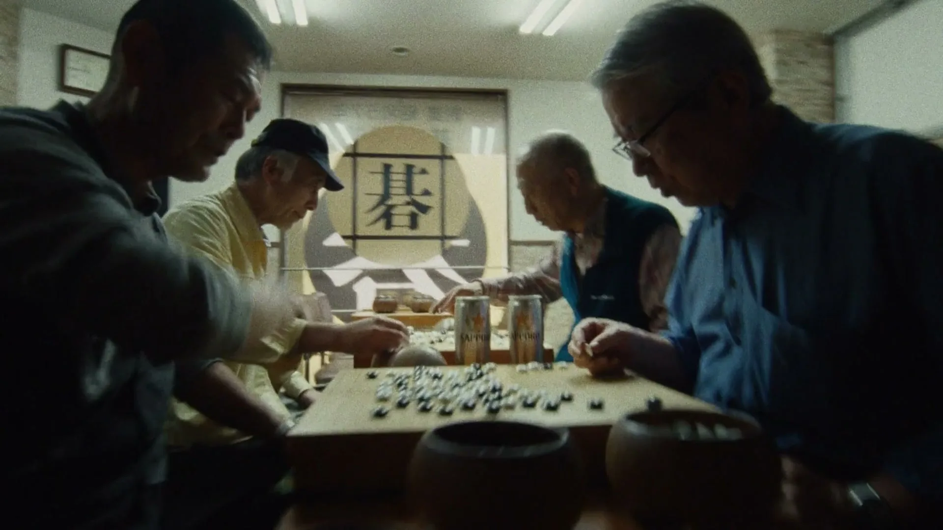 Four older men play a game of Go around a wooden table in a brightly lit room, with Sapporo cans visible.