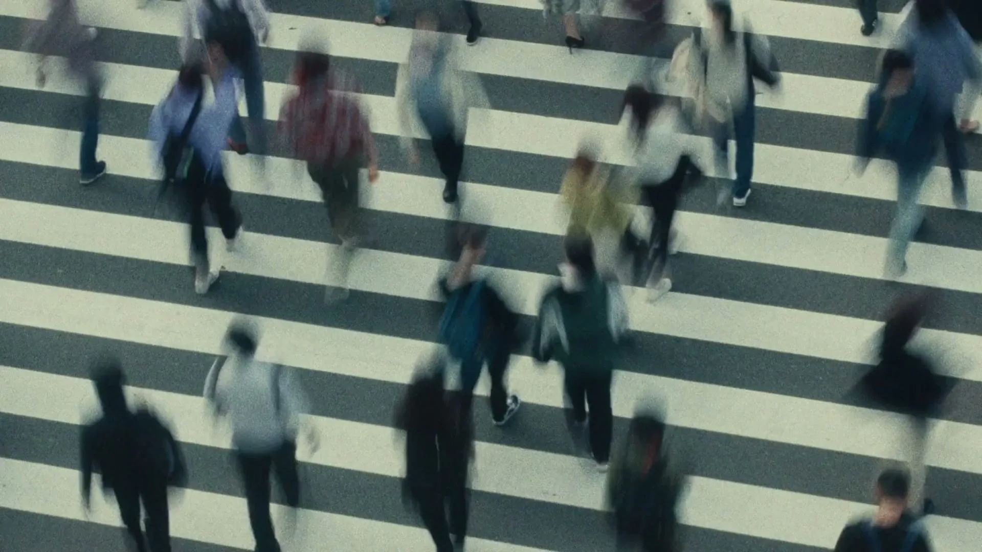 An overhead shot shows a crowd of people crossing a busy street at a crosswalk, their movements blurred by motion.
