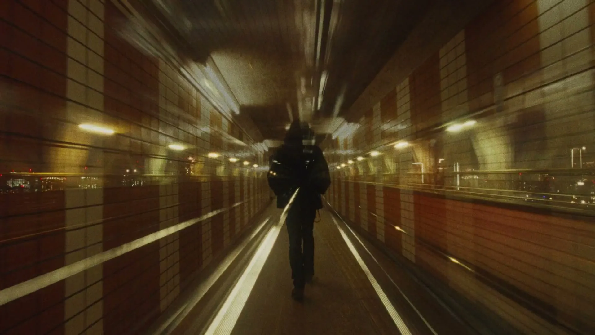 A person with long dark hair walks away from the camera down a brightly lit tunnel with orange and white striped walls.