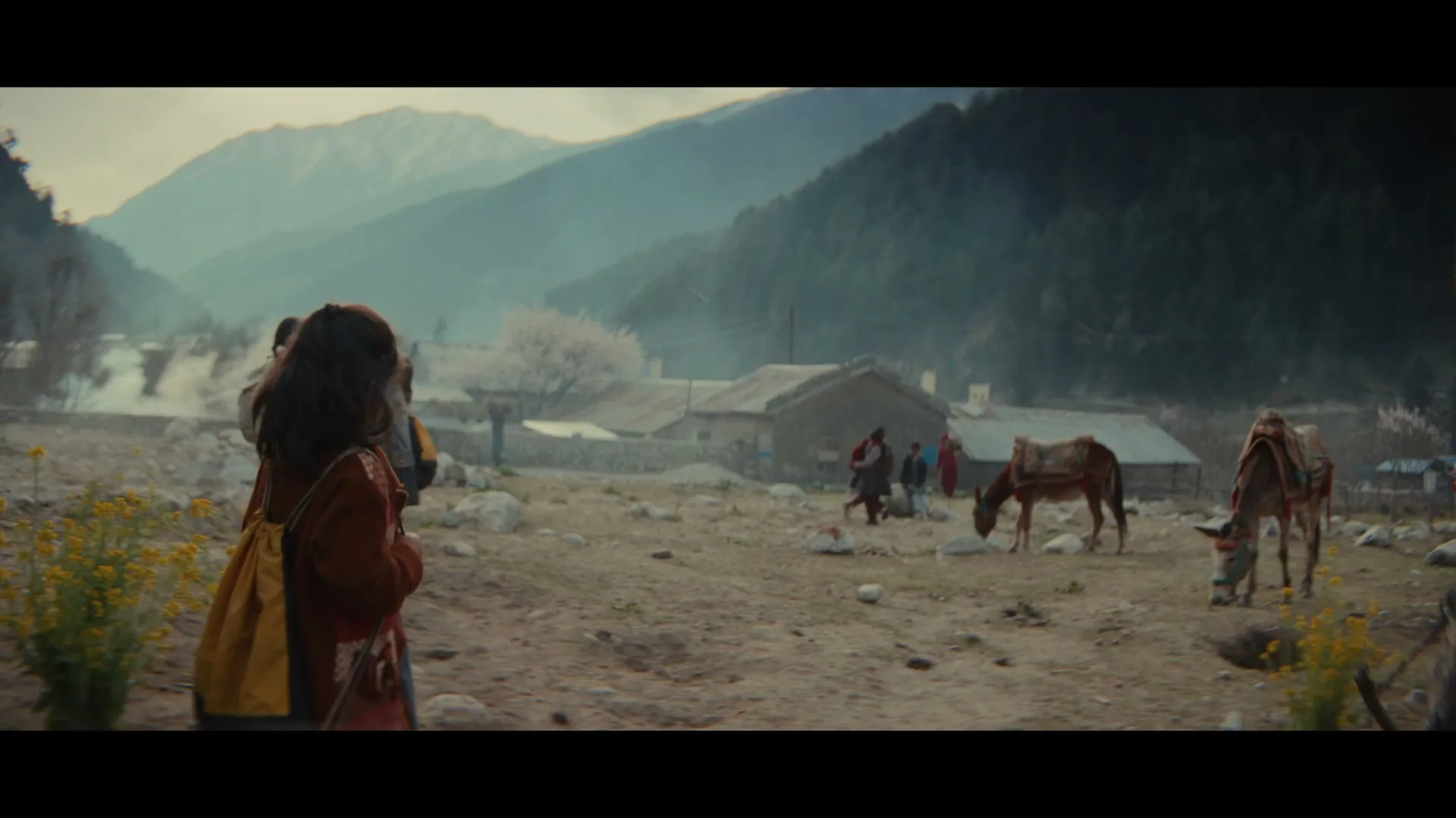 A young girl with a yellow backpack smiles at the camera, then turns and runs towards other children in a rural village with mountains in the background.