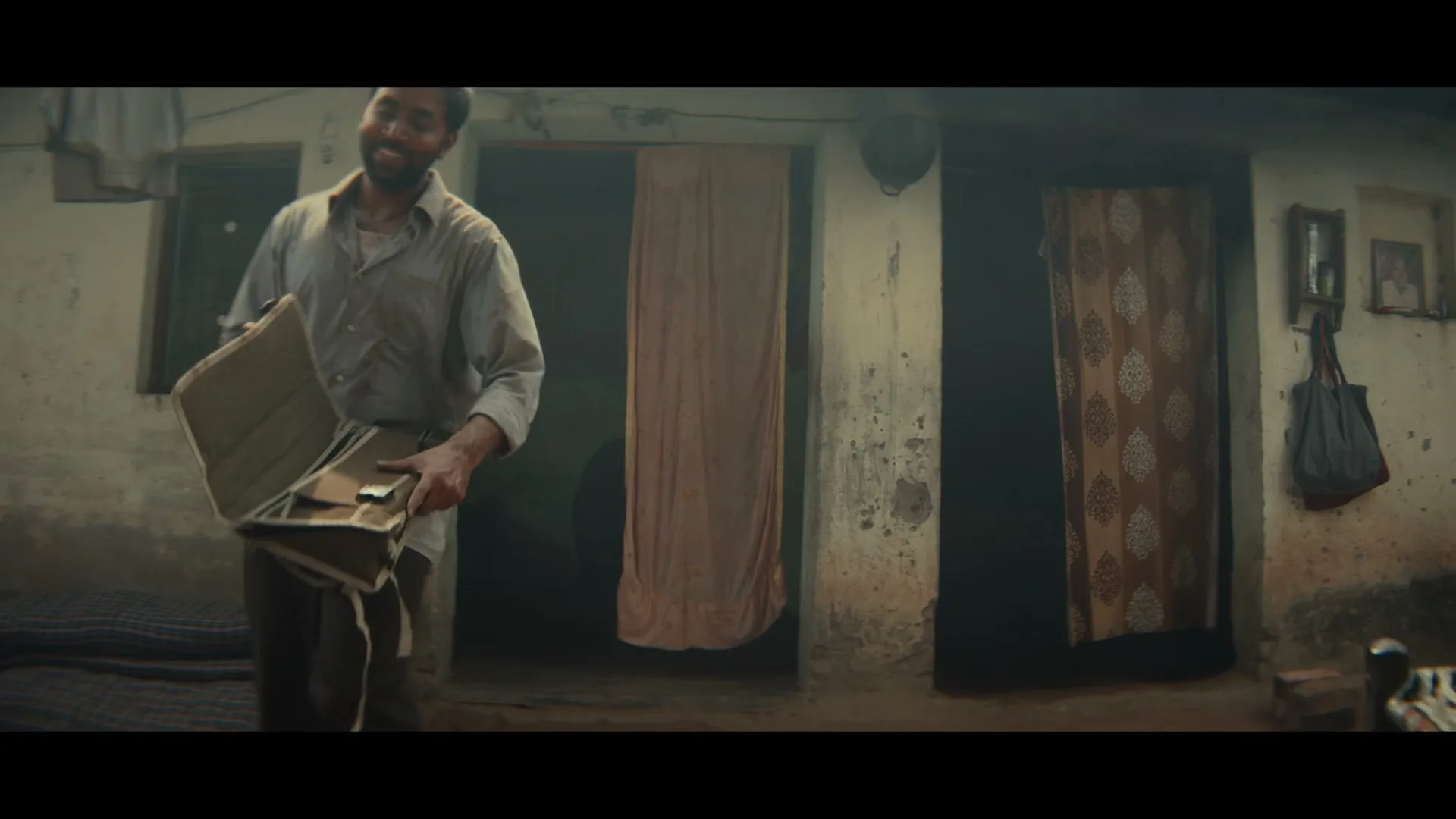 A man with a beard, wearing a light shirt and dark pants, walks out of a doorway holding a portable sewing machine, then bends down to place it on a mat outside a rustic home.