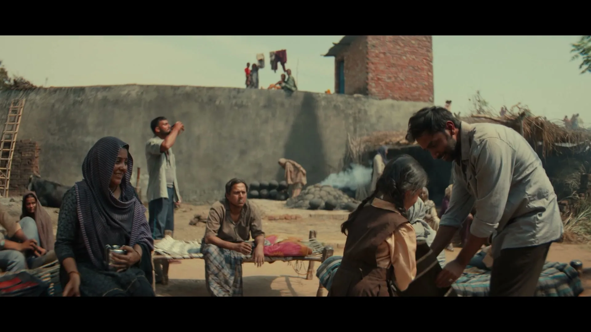 A man in a light shirt gives a small girl a bowl while other villagers sit around them in a rural outdoor setting.