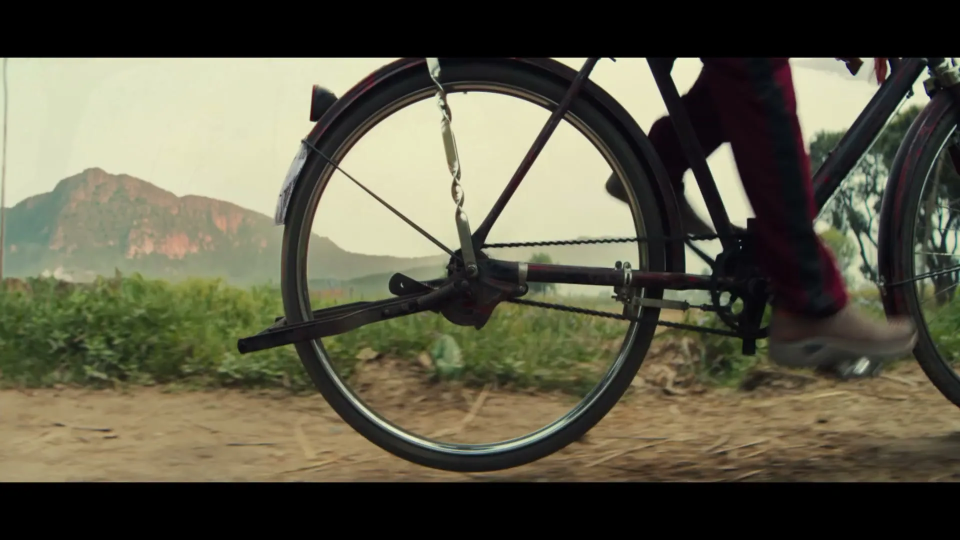 A young girl walks past a man operating a large circular farm machine, then a close-up of a bicycle wheel on a dirt path with mountains in the background.