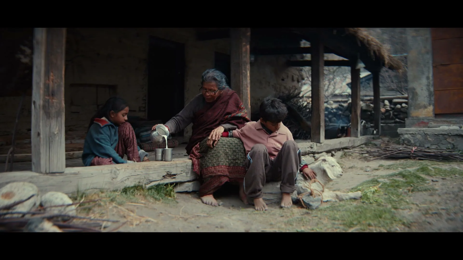 An elderly woman pours milk into a cup for a young boy and girl sitting on a porch with a goat in a rural village.