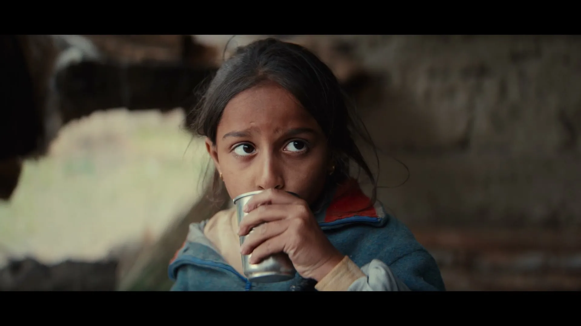 A young girl with dark hair and a blue jacket drinks from a metal cup, looking up and to the right with a thoughtful expression in an interior setting.