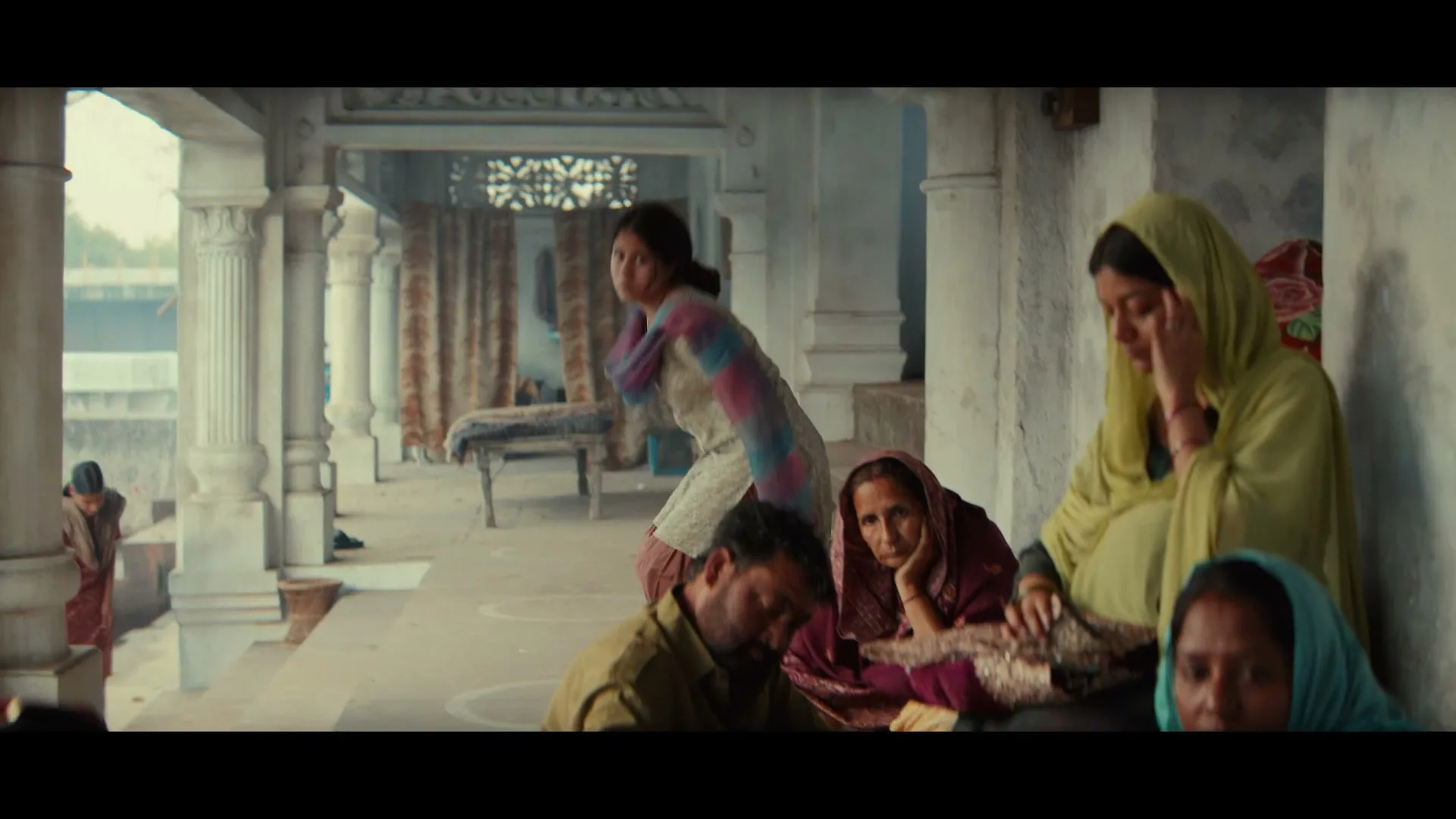 A young woman in traditional Indian attire stands up and walks away from a group of people sitting on a stone porch in a rural setting.